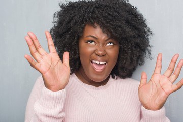 Wall Mural - Young african american plus size woman over grey grunge wall wearing winter sweater crazy and mad shouting and yelling with aggressive expression and arms raised. Frustration concept.