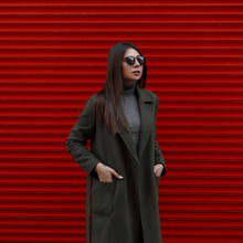 Fashion Young Girl With Stylish Sunglasses In A Fashionable Green Coat Stands Near The Red Metal Wall