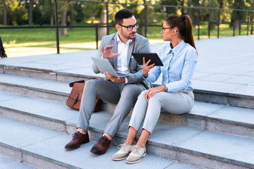 Team of two business people working together outdoors using laptop and tablet, discussing business ideas and planning future projects.