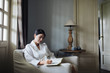 © LUMINA IMAGES - Young woman writing in notebook while sitting on armchair at home