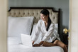 © LUMINA IMAGES - Beautiful Asian woman sitting on a king-size bed and typing on her laptop.