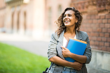 pretty cheerful latin american student smiling at camera carrying notebook on campus at college