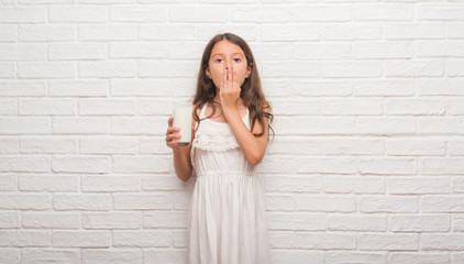 Canvas Print - Young hispanic kid over white brick wall drinking a glass of milk cover mouth with hand shocked with shame for mistake, expression of fear, scared in silence, secret concept