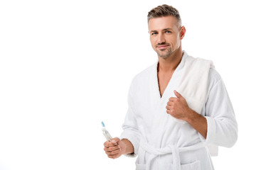 happy adult man in bathrobe with towel over shoulder holding toothpaste and toothbrush isolated on white