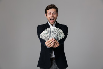 Poster - Image of joyful businessman 30s in suit smiling and holding fan of money in dollar banknotes, isolated over gray background