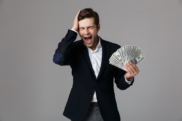 Canvas Print - Image of excited businessman 30s in suit smiling and holding fan of money in dollar banknotes, isolated over gray background