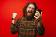 © Vulp - Amazed bearded hipster man wearing round eyeglasses holding smartphone and celebrating success, victory over red background