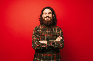 portrait of cheerful bearded hipster man with crossed arms over red background