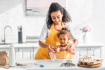 smiling african american mother helping daughter breaking egg for preparing dough in kitchen