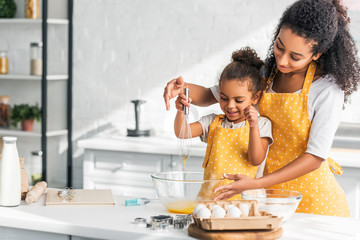 african american mother and daughter whisking eggs for dough in kitchen