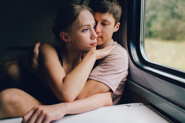 Mood portrait of young lifestyle couple of lovers traveling in train. Beautiful loving pair hugging, kissing, telling secret near window. Boy and girl in love.  Friends in journey on vacation.