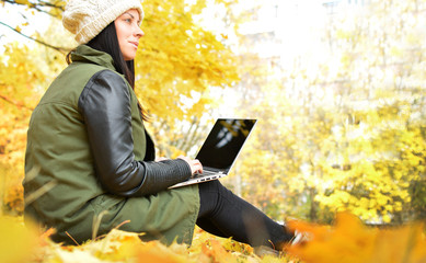 Wall Mural - Girl in hipster with laptop in autumn park. A woman in a cap using a laptop while sitting on fallen leaves. Freelancer in the hat uses remote communication technology. Remote work. Free space