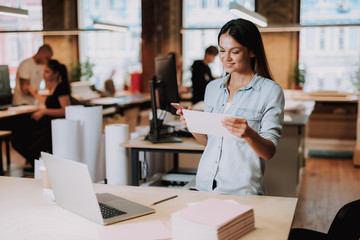 Waist up portrait of charming young lady standing near table with laptop and holding smartphone. She is smiling