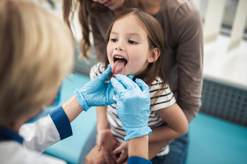 Wall Mural - Concept of professional consultation and examination. Close up portrait of little girl with opening mouth being examined by pediatrician woman in medical office