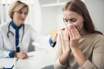 Wall Mural - Concept of professional consultation and disease. Waist up portrait of young female patient feeling bad and blowing nose in medical office