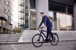 © HD92 - one young man, 20-29 years old, wearing suit, looking smiling. riding, pedaling standing, fancy  bicycle. full length body. modern architecture building behind.