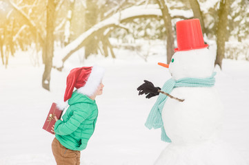 Wall Mural - Little boy in red christmas hat holds gift box for snowman