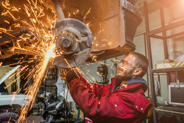 Young caucasian man in red work clothing repairing car with angle cutter. Many sparks on background.