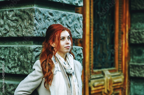 Fényképezés Portrait of a young beautiful fashionable red-haired woman with long curly hair , posing on the streets of the European city
