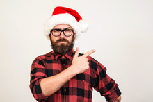 Handsome Bearded Man In Santa Hat And Glasses. Santa Man Pointing To The Side Over White Wall. Christmas Concept. Mock Up, Copy Space For Text. Hipster Bearded Man Thinking For Christmas Gift Ideas.