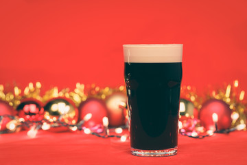 Dark ale beer in pint glass with christmas baubles tinsel and lights on red background toned