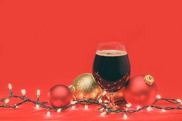 Dark ale beer in a tulip glass with christmas baubles and lights on red background toned