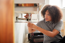 Young Black Female Plumber Sitting On The Floor Fixing A Bathroom Sink, Seen From Doorway