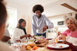 © Monkey Business - Middle aged black man carving and serving meat at Sunday family dinner with his partner, kids and their grandparents, front view