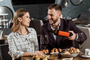 Happy young adult couple having dinner while man pouring wine in glasses in restaurant