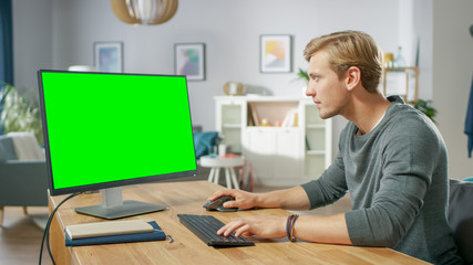Handsome Young Man Works on a Green Mock-up Screen Personal Computer while Sitting at His Desk in the Cozy Apartment.