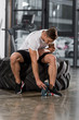 © LIGHTFIELD STUDIOS - handsome athletic bodybuilder sitting on tire and taking sport bottle of water in gym