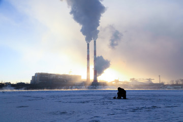 Wall Mural - Winter fishing, men's passion, fishermen catch fish on a frozen river against the background of the factory pipes at sunset. Concept of oil gas processing plants, coal mining and minerals