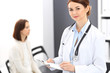 © rogerphoto - Doctor woman at work. Portrait of female physician filling up medical form while standing near reception desk at clinic or emergency hospital. Patient woman sitting at the background. Medicine concept