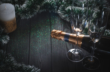 A bottle of champagne and two glasses stand on a dark gray wooden table surrounded by Christmas decorations