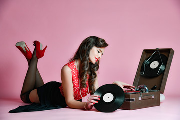 Glamorous pinup girl lying near retro gramophone holding LP vinyl record looking up on pink background