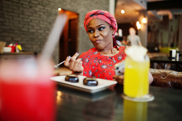 Wall Mural - Stylish african woman in red shirt and hat posed indoor cafe, eat chocolate dessert cakes and looking at mobile phone.