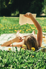 Wall Mural - Young woman lying down on a blanket in home yard and sunbathing on a sunny day and reading a real book. Grass and dandilion background, nice vintage look.