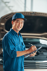 Wall Mural - Portrait of young auto mechanic in uniform standing and writing a conclusion after inspection of a car at car service