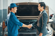 Asian young auto mechanic in uniform and a client are shaking hands and smiling with car in the background