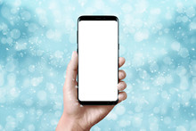 Modern Black Phone In Hand. Isolated, Blank Screen For Mockup. Christmas Scene With Blue Background With Snowflakes, Light And Bokeh.