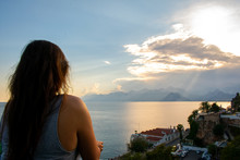 Beautiful Young Woman With Dark Hair Standing On Balcony And Enjoying The View. Sunset Over Mountains And Sea In Antalya, Turkey 