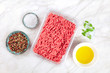 © laplateresca - A photo of minced meat in a plastic tray, shot from above on a white marble background with salt, pepper, olive oil, parsley leaves and copy space