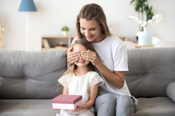 Loving young mother closing eyes of little smiling daughter making surprise to happy kid girl holding gift box sitting on sofa at home, excited child receiving birthday present from caring mom