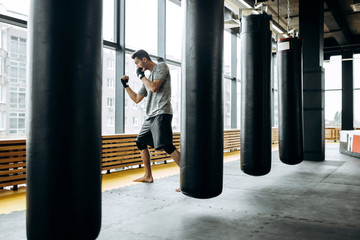 Dark-haired guy dressed in the grey t-shirt and black shorts stands behind the hanging punching bag next to panoramic windows in the boxing gym