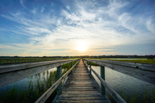 Coastal Waters With A Very Long Wooden Boardwalk Pier In The Center During A Colorful Summer Sunset Under An Expressive Sky With Reflections In The Water And Marsh Grass In The Foreground