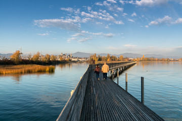 Canvas Print - landscape view of marsh and lake shore with the town of Rapperswil in evening light and a long wooden boardwalk in the foreground and a couple going for a leisurely walk