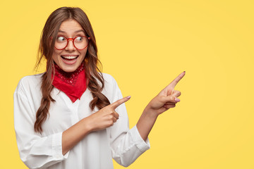 Wall Mural - Studio shot of content woman with European appearance, has joyful expression, indicates with both index fingers, shows free space for your advertising content. Joyful girl in white shirt and bandana