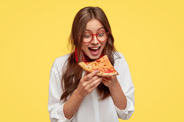 Hungry student opens mouth widely while sees delicious slice of pizza, wants to eat, dressed in white shirt, models against yellow background. Positive woman with junk food. People and eating