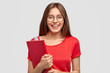 © wayhome.studio  - Positive Caucasian girl with charming smile, wears red t shirt, holds textbook, models against white background, has mood for studying, wears optical glasses for good vision. Youth, learning concept