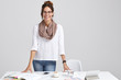 Satisfied intelligent female tutor wears white jumper and jeans, keeps hands on table, ready to give private lessons, drinks coffee, stands against white background. Woman works freelance at project